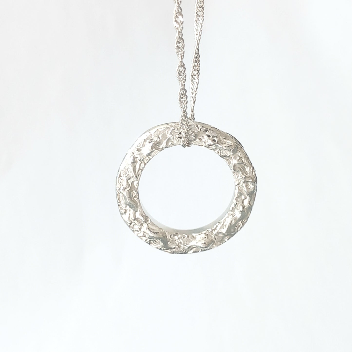 Textured circle silver pendant with a wave like raised silver texture. A chain threaded through the top of the circle on a white background.