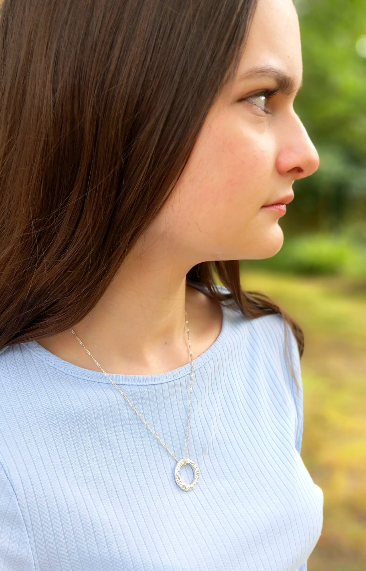 A dark haired lady with a blue top wearing a silver textured circle pendant where the chain is threaded through the top. With a green background.