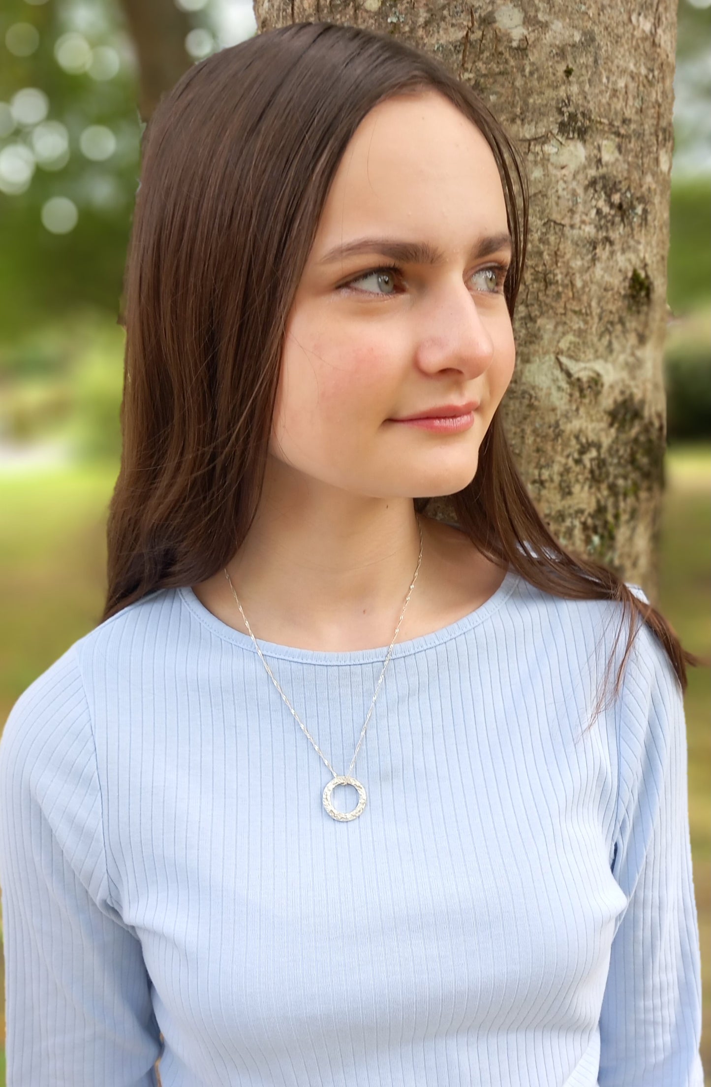 A dark haired lady wearing a silver textured circle pendant where the chain is threaded through the top