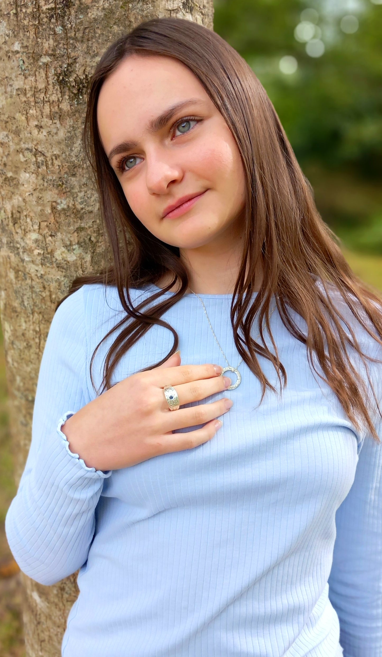 A dark hairded lady leaning against a tree looking into the distance, wearing a silver textured circle pendant where the chain is threaded through the top. A silver sapphire raised textured ring is on her finger.