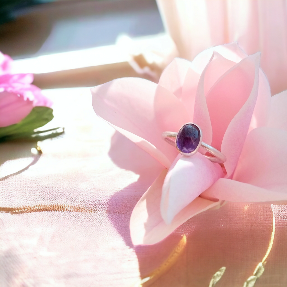An amethyst cabochon cut stone in a simple solid silver ring nestled in a pink flower in sunlight on a wooden window sill.