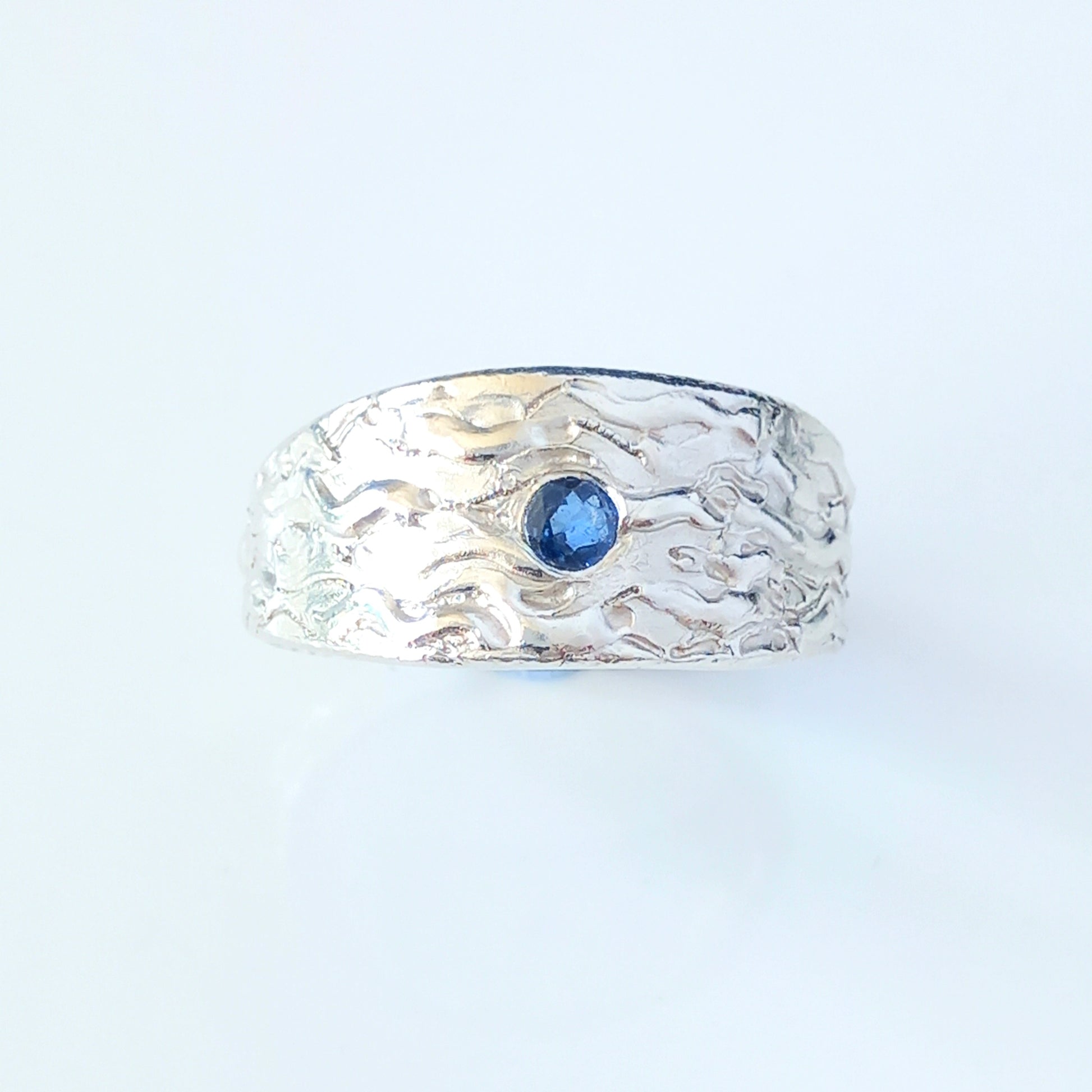 Thick silver ring with a wave texture and a round blue sapphire in the centre. On a white shiny surface.