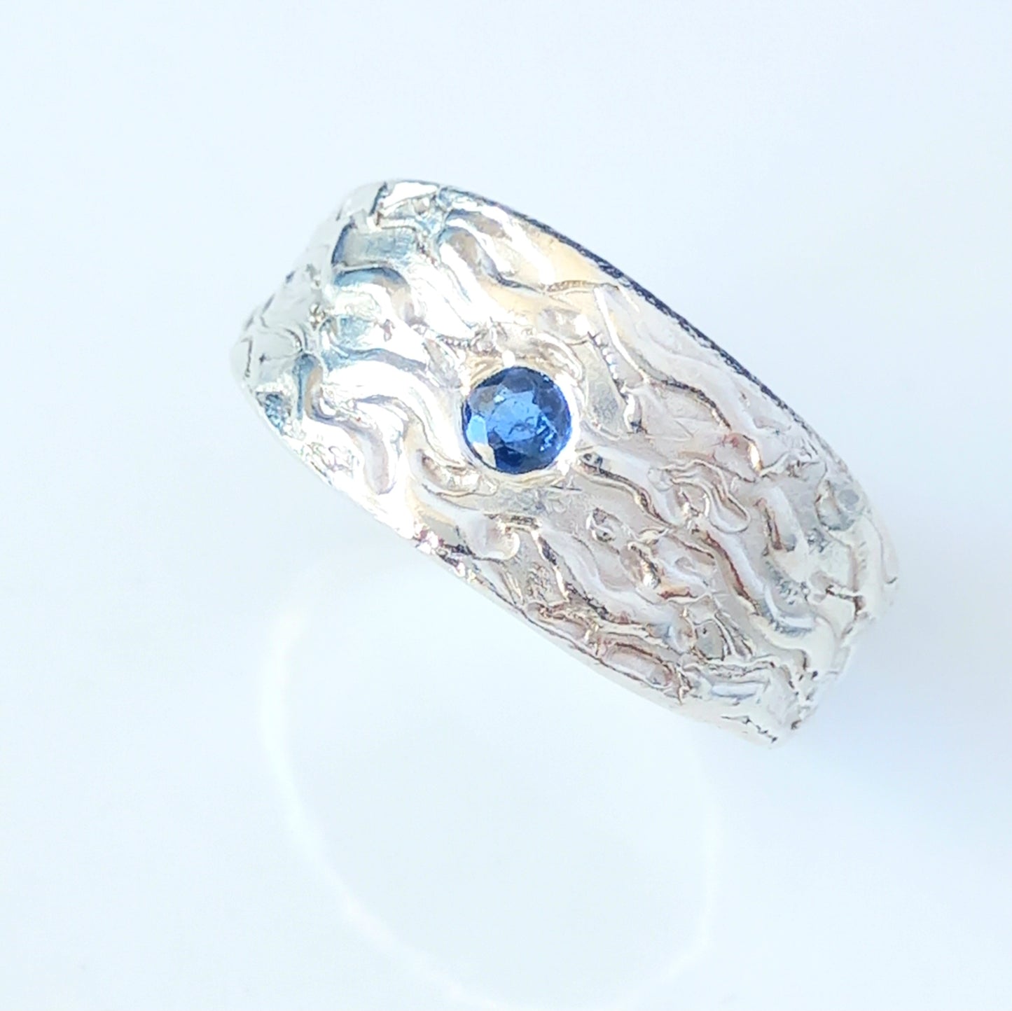 Thick silver ring with a wave texture and a round blue sapphire in the centre. On a white shiny surface.