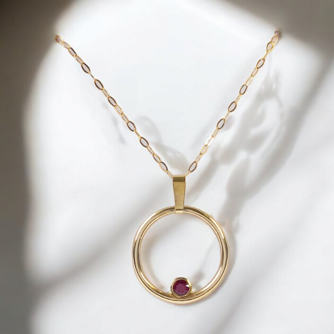 A gold circle pendant with a ruby set inside the circle. With a chain through the bail and a white background with shadows.