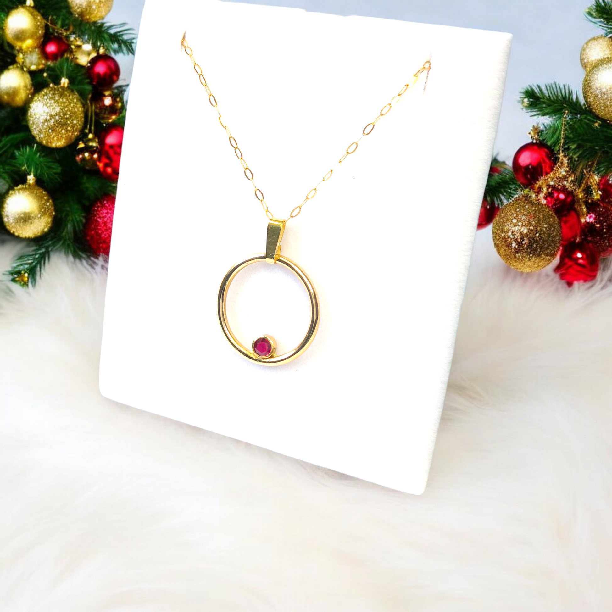 A gold circle pendant with a ruby set inside the circle. On a jewellery stand with red and gold Christmas decorations.