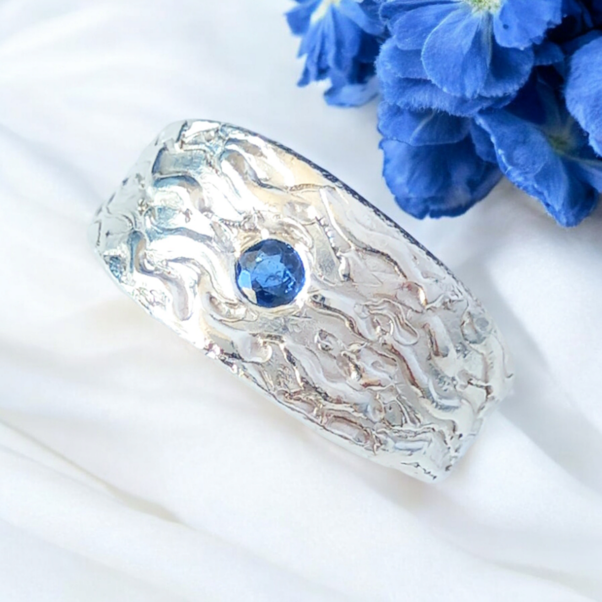 Thick silver ring with a wave texture and a round blue sapphire in the centre. On a white shiny fabric and a blue flower to the side.
