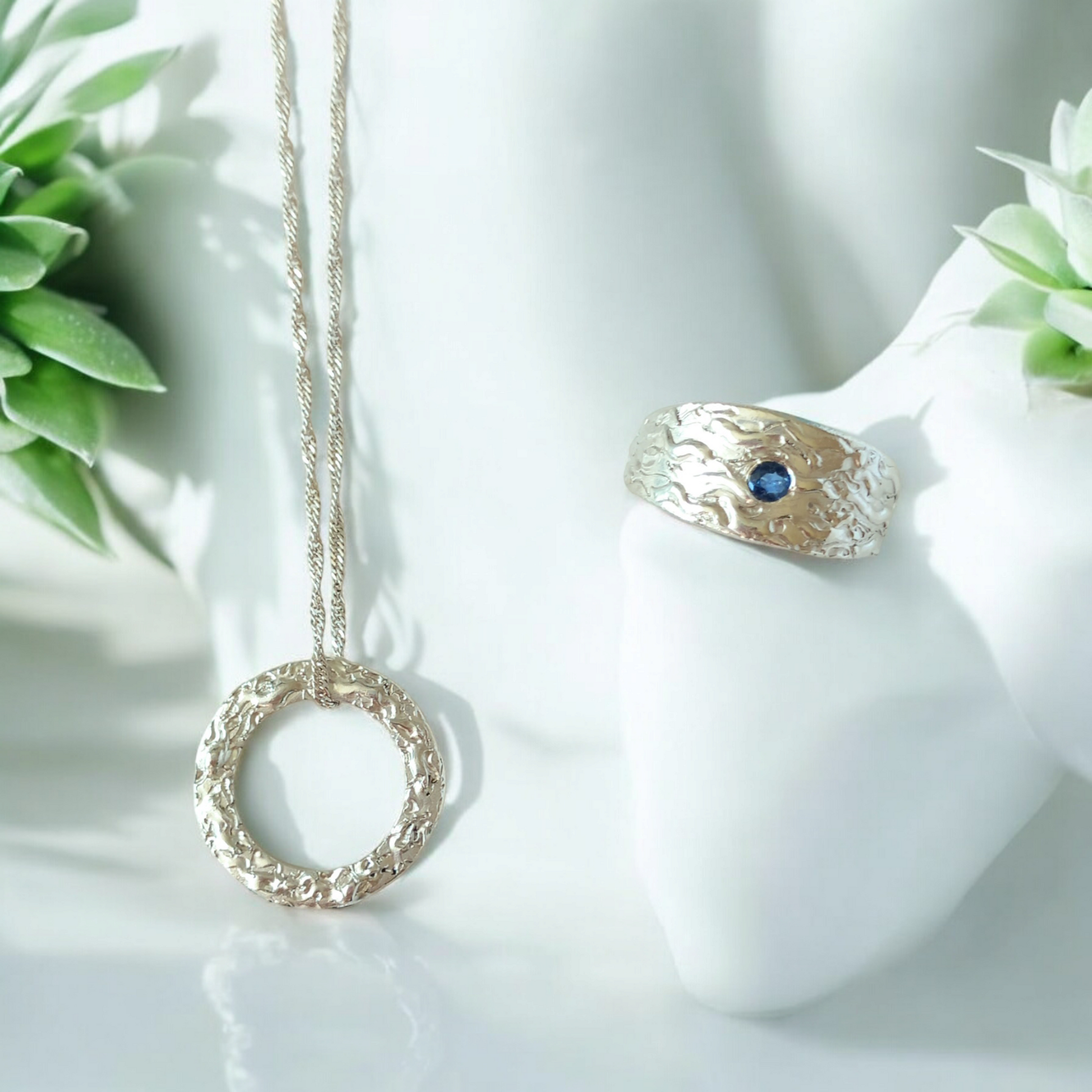 A silver textured circle pendant where the chain is threaded through the top leaning against a white background with a green plant in the background. Also a chunky sapphire wave textured ring resting at an angle with another green plant to the right.