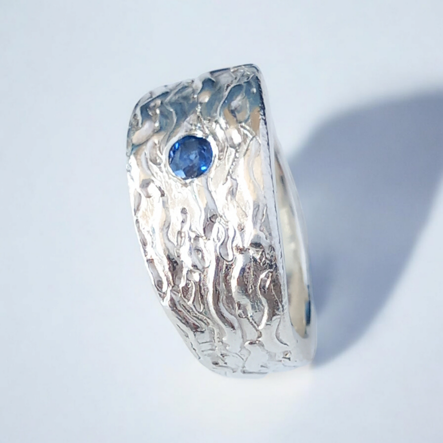 Thick silver ring with a wave texture and a round blue sapphire in the centre. On a white shiny surface.