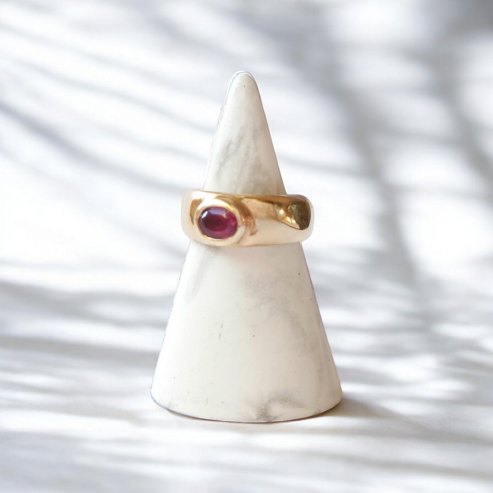 A thick gold Roman style ring with an oval pink garnet stone on a marble stand. Set on a white marble countertop with sunlight and shadows falling across the ring.
