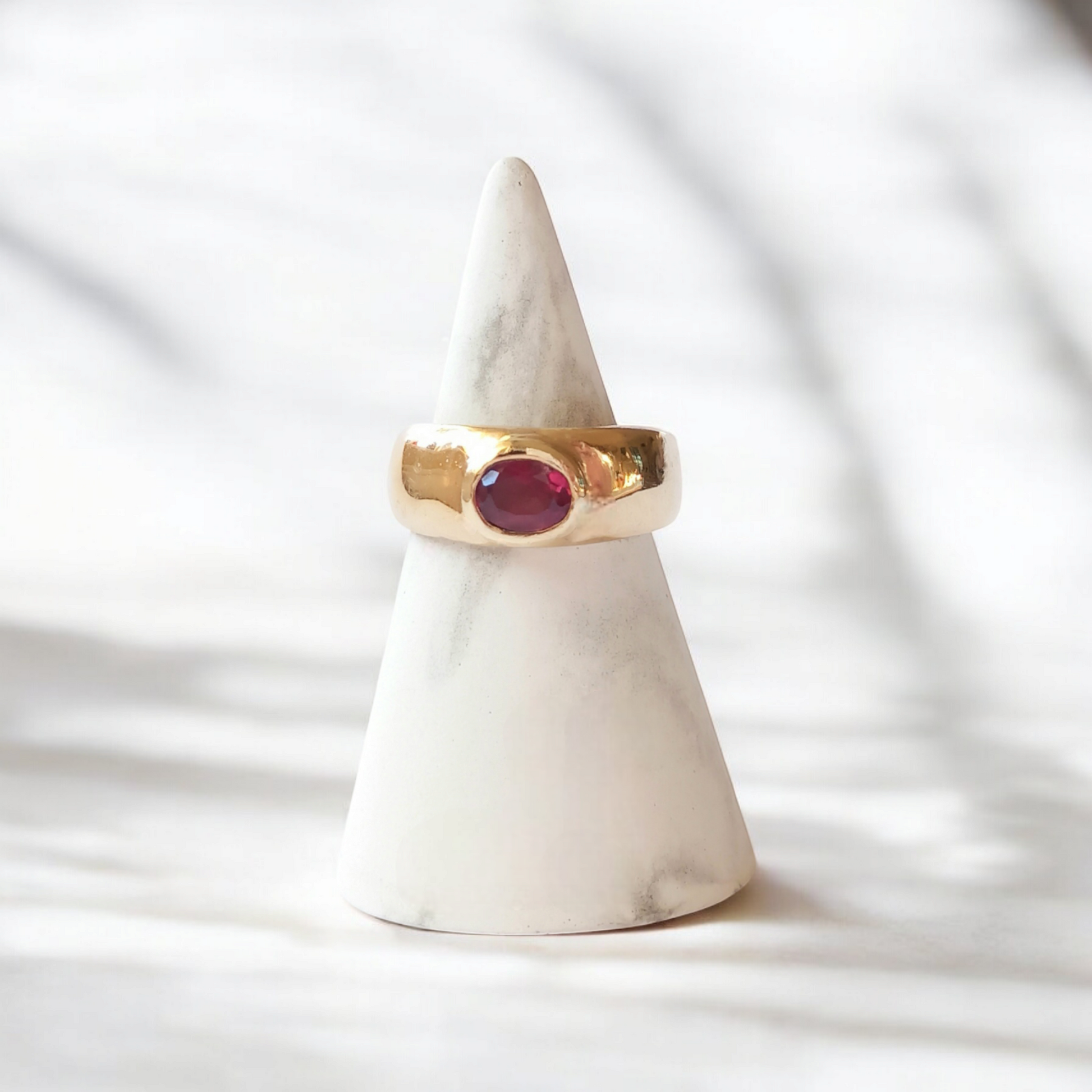 A thick gold Roman style ring with an oval pink garnet stone on a marble stand. Set on a white marble countertop with sunlight and shadows falling across the ring.