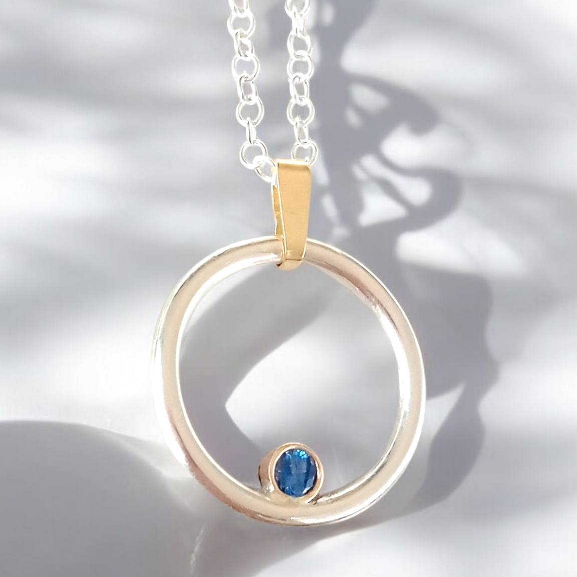 A silver and gold round pendant with a round sapphire set inside. On a silver chain with light and shadows across the pendant.