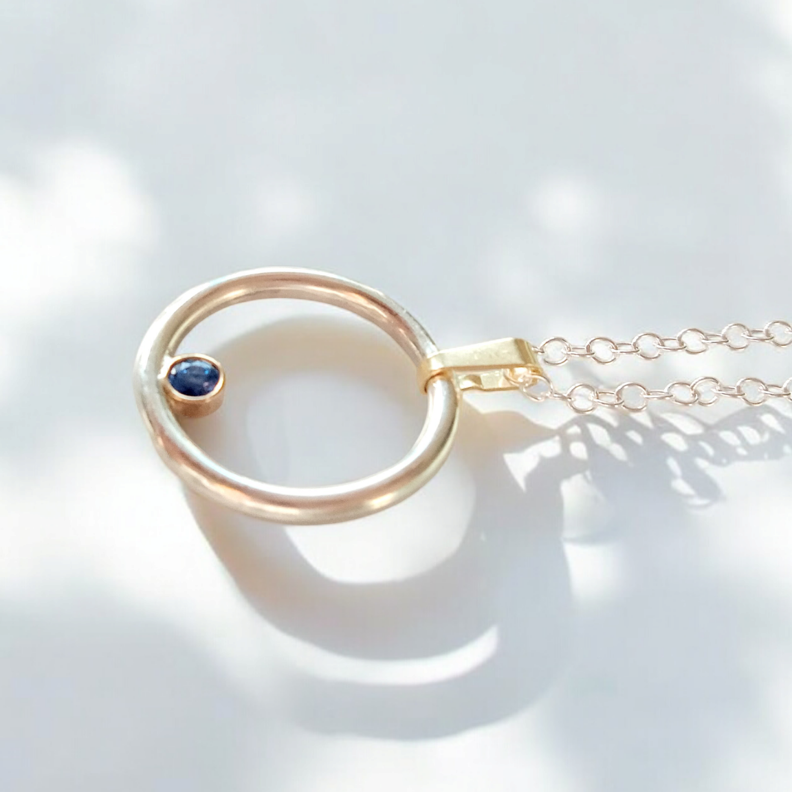 A silver and gold round pendant with a round sapphire set inside. On a silver chain with light and shadows across the pendant.