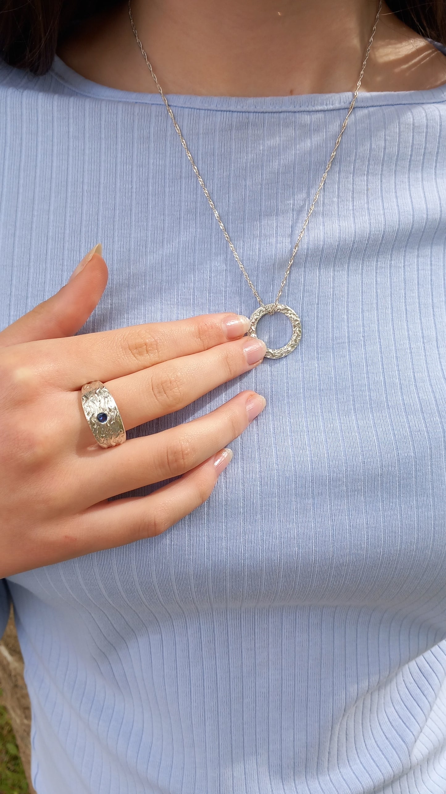 Thick silver ring with a wave texture and a round blue sapphire in the centre. On a hand pointing towards a matching circle textured with waves.
