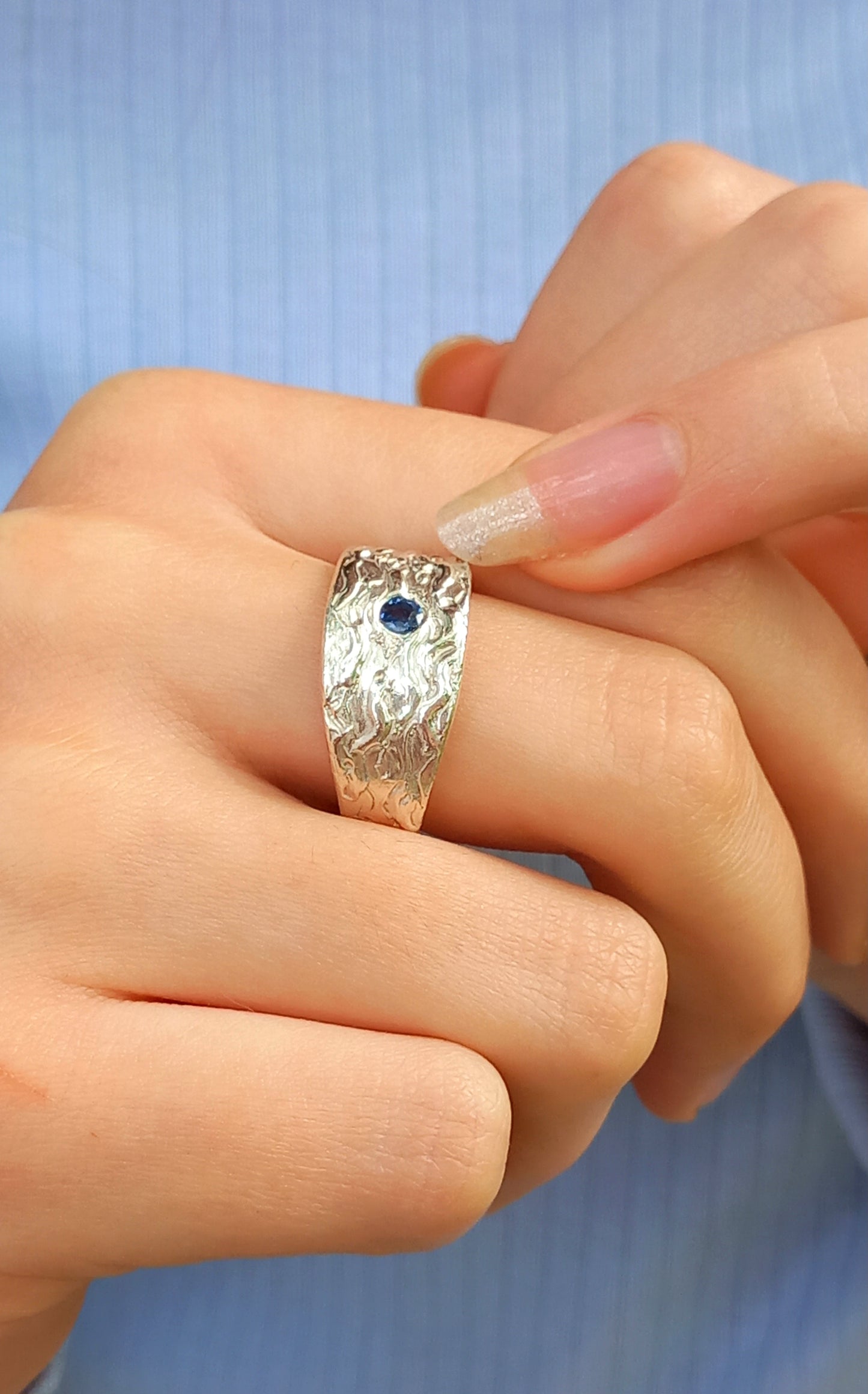 Thick silver ring with a wave texture and a round blue sapphire in the centre. On a hand with a long fingernail pointing towards the ring.
