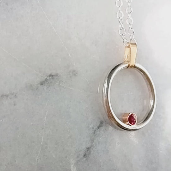 Silver and gold circle pendant with a ruby in a gold setting nestled inside the silver circle.  A clip on gold bail at the top on a silver chain.  Video starts at the side and pans over to the other with a close up of the ruby.  All on a grey and white marble shiny background.