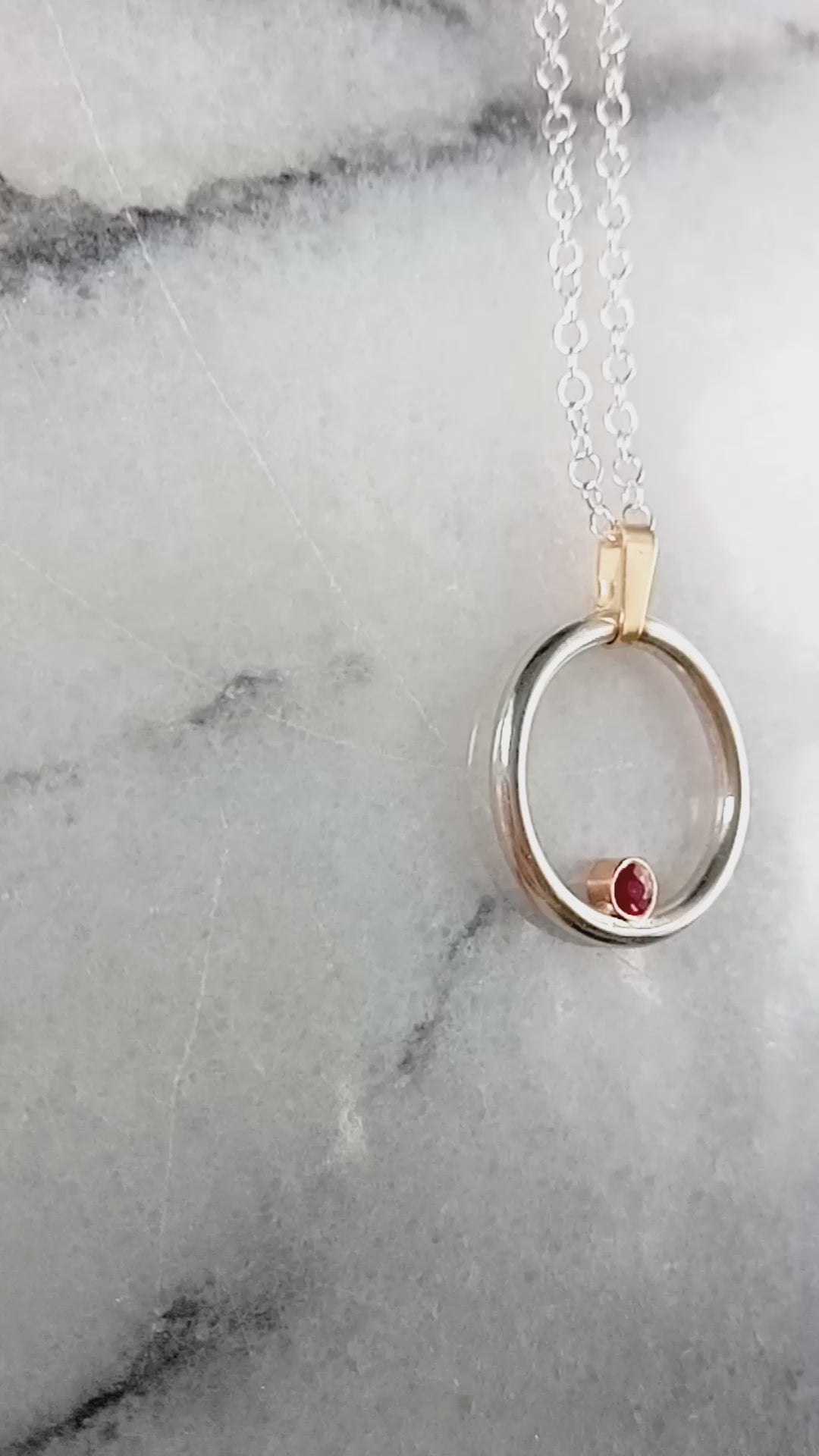 Silver and gold circle pendant with a ruby in a gold setting nestled inside the silver circle.  A clip on gold bail at the top on a silver chain.  Video starts at the side and pans over to the other with a close up of the ruby.  All on a grey and white marble shiny background.