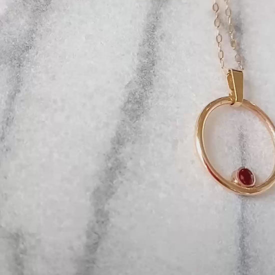 video of gold circle pendant on a marble background.  Within the circle pendant a round bezel set ruby is nestled in the 6 O'clock position.