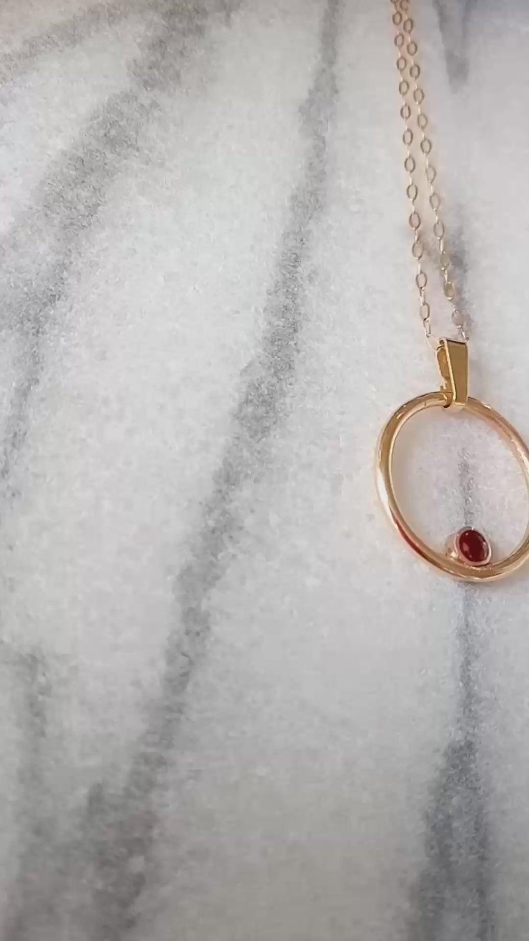 video of gold circle pendant on a marble background.  Within the circle pendant a round bezel set ruby is nestled in the 6 O'clock position.